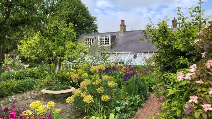 Monk's House and conservatory viewed through the colourful florals in the cottage garden. There is a blue sky with white clouds
